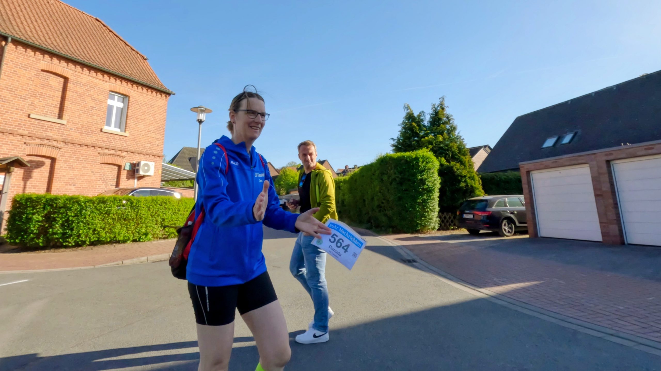 Two runners participating in a street race, smiling as they pass a residential area with brick houses on a sunny day.