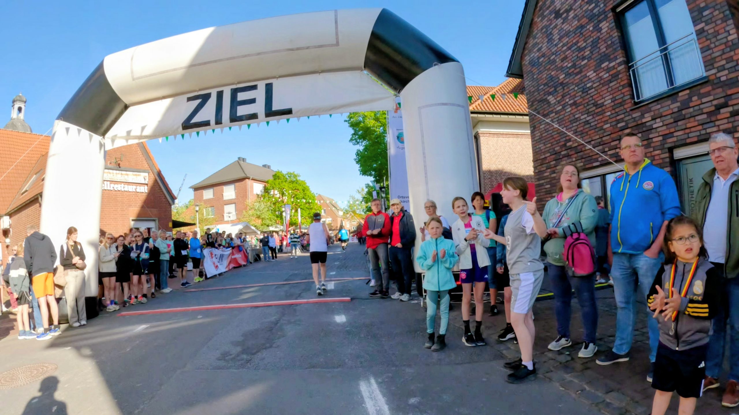 Crowd of spectators cheering as a runner crosses the finish line beneath a white inflatable arch marked 'ZIEL' in a small-town street.