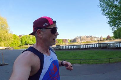 Runner in a white and blue singlet wearing a pink and black cap and sunglasses runs along a riverside park with trees and a balustrade in the background