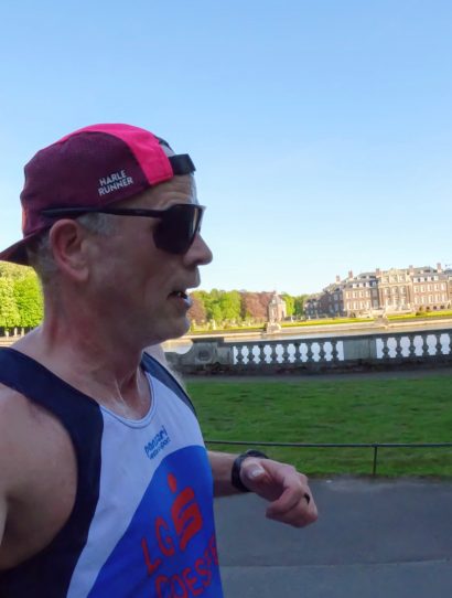 Runner in a white and blue singlet wearing a pink and black cap and sunglasses runs along a riverside park with trees and a balustrade in the background