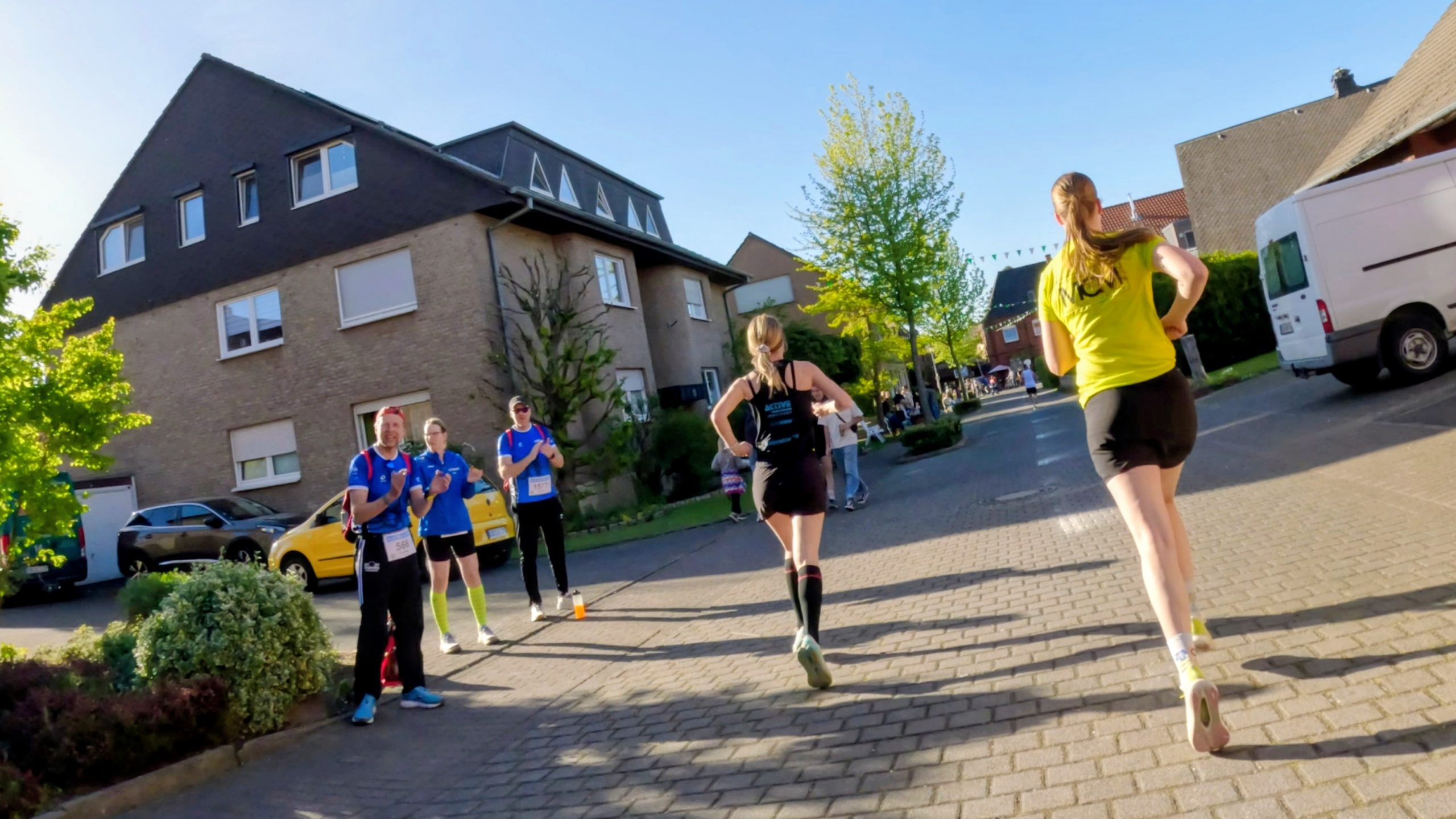 Runners in blue shirts walking/standing on a suburban street during a sunny race, houses and trees lining the block