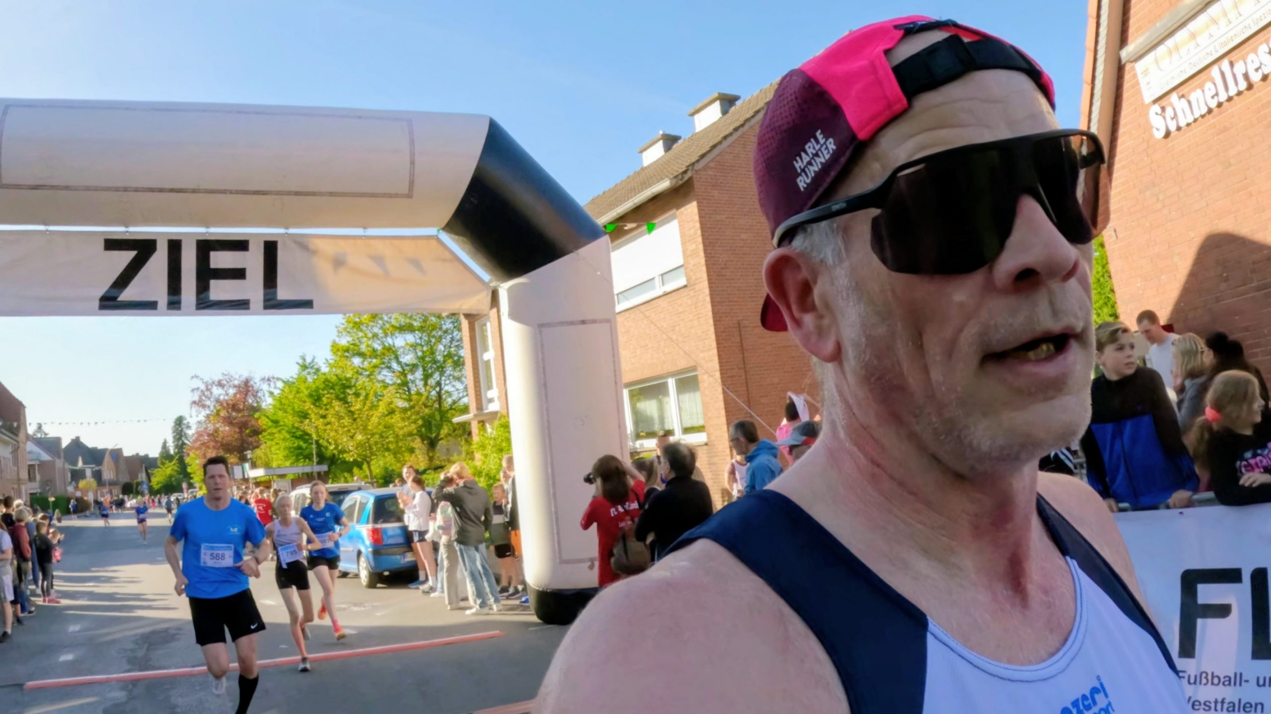Close-up of a male runner wearing a red cap and large sunglasses at the finish line, with other runners and spectators behind him.