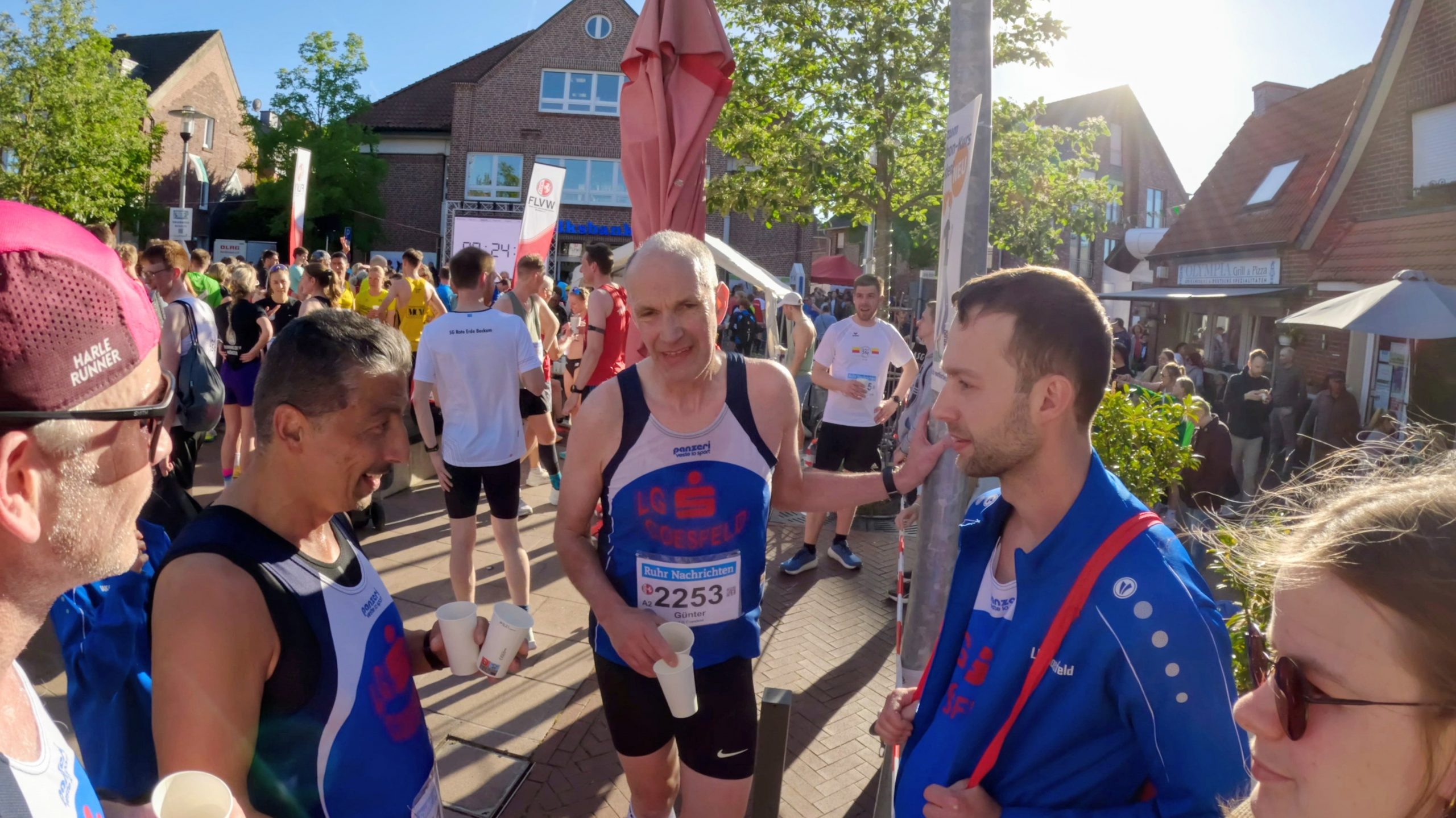 Group of marathon runners in colorful race bibs and blue uniforms chat and stretch before a race in a sunny urban street.