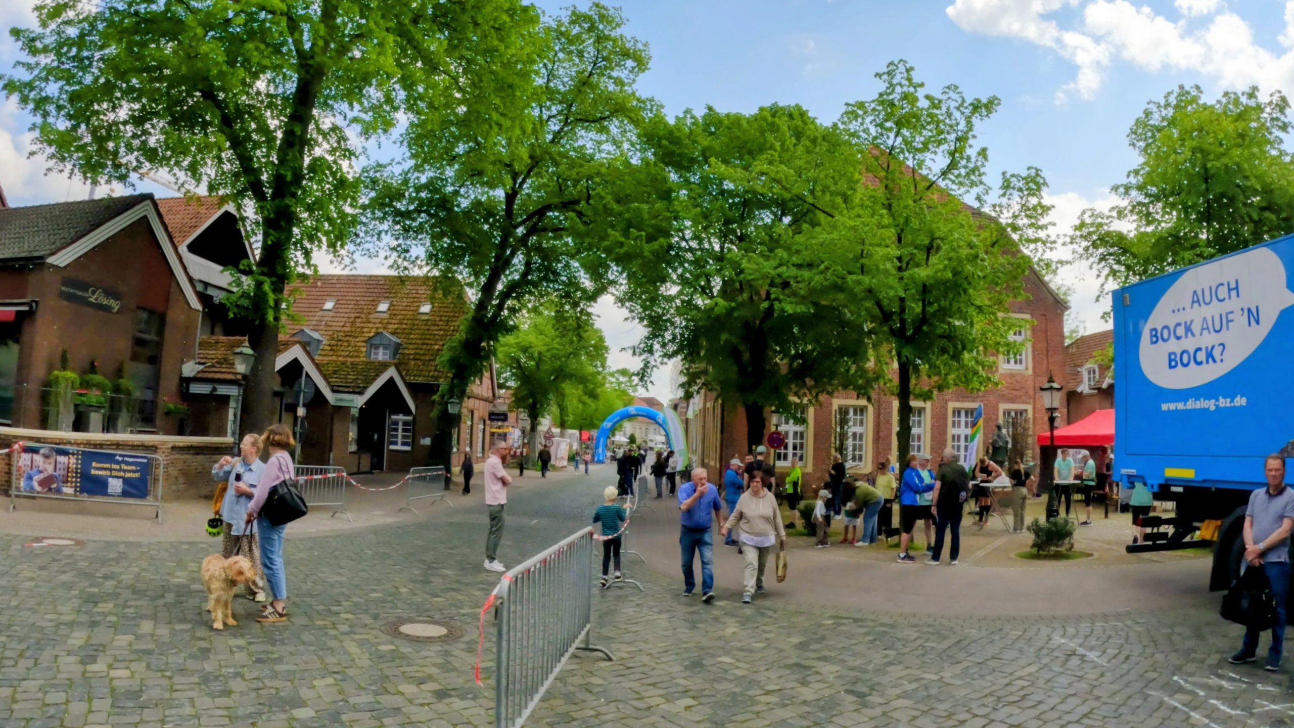 Busy town street at a fair: people stroll among trees, cobblestones, and colorful storefronts with a large blue promotional truck that reads '... AUCH BOCK AUF'N BOCK?'