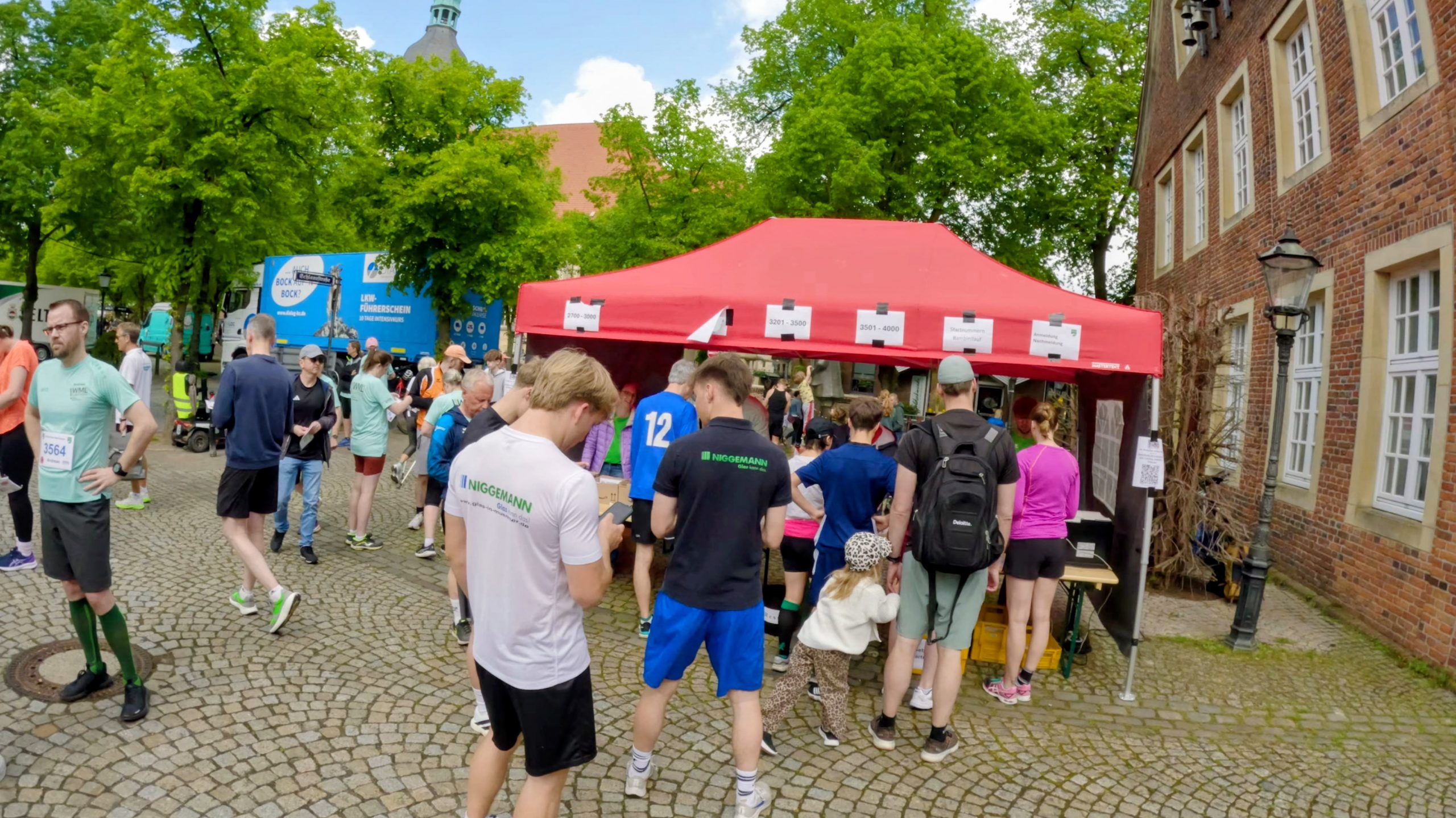 Crowd of runners at a race checkpoint under a red canopy, with a brick building and green trees in the background.