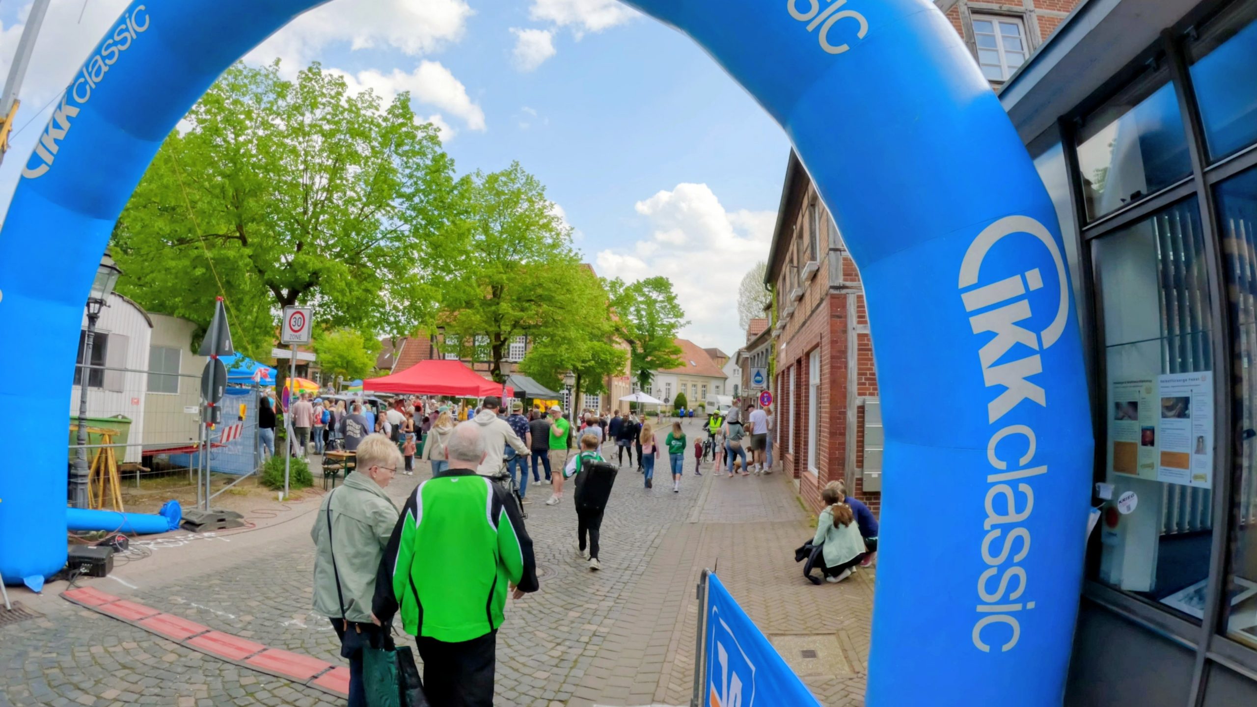 Crowd of people at a street festival with red and white tents under a bright blue inflatable archway.