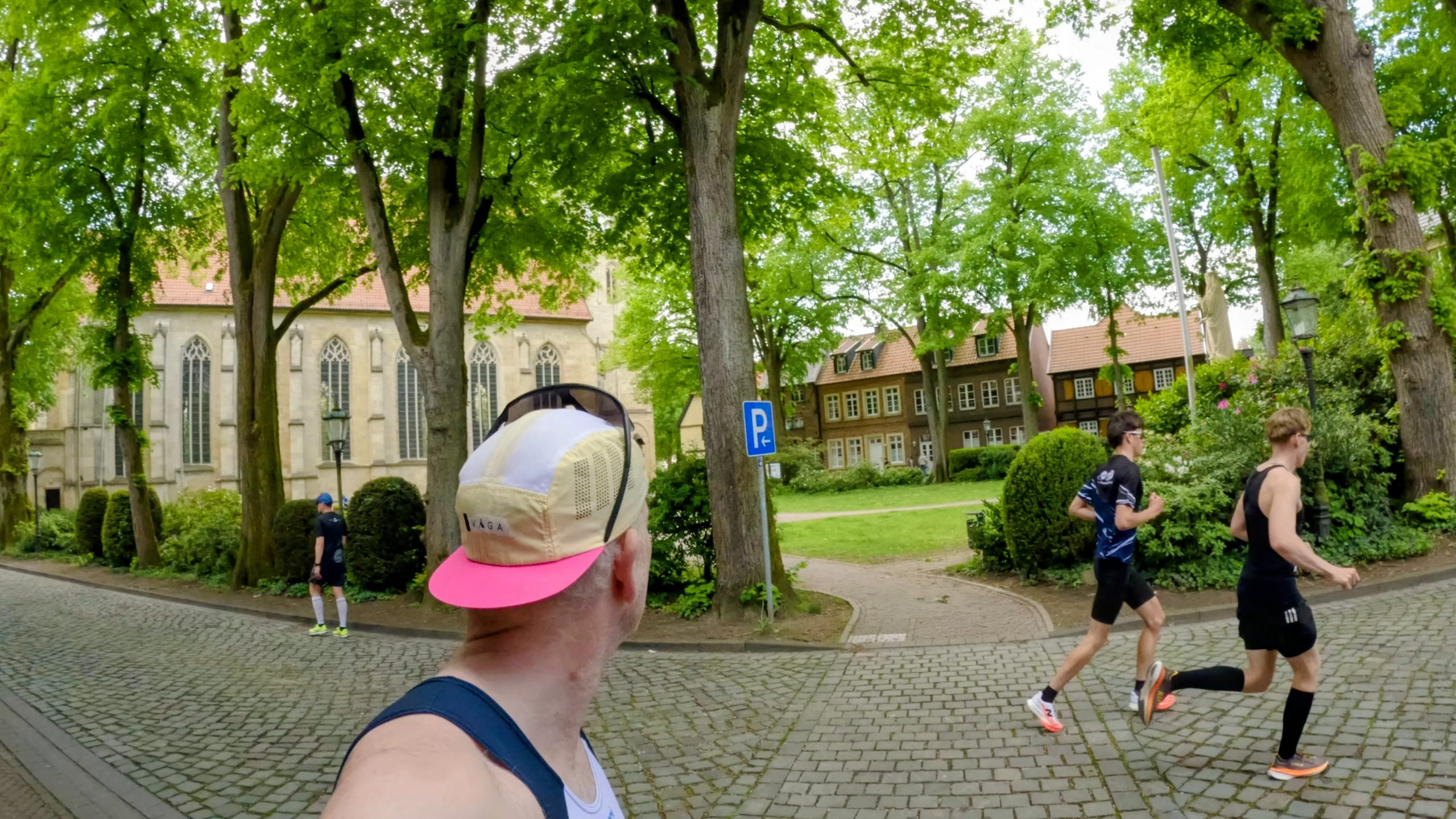 Runners pass a church and leafy trees along a sunlit cobblestone street; foreground runner wears a beige cap with a pink bill.