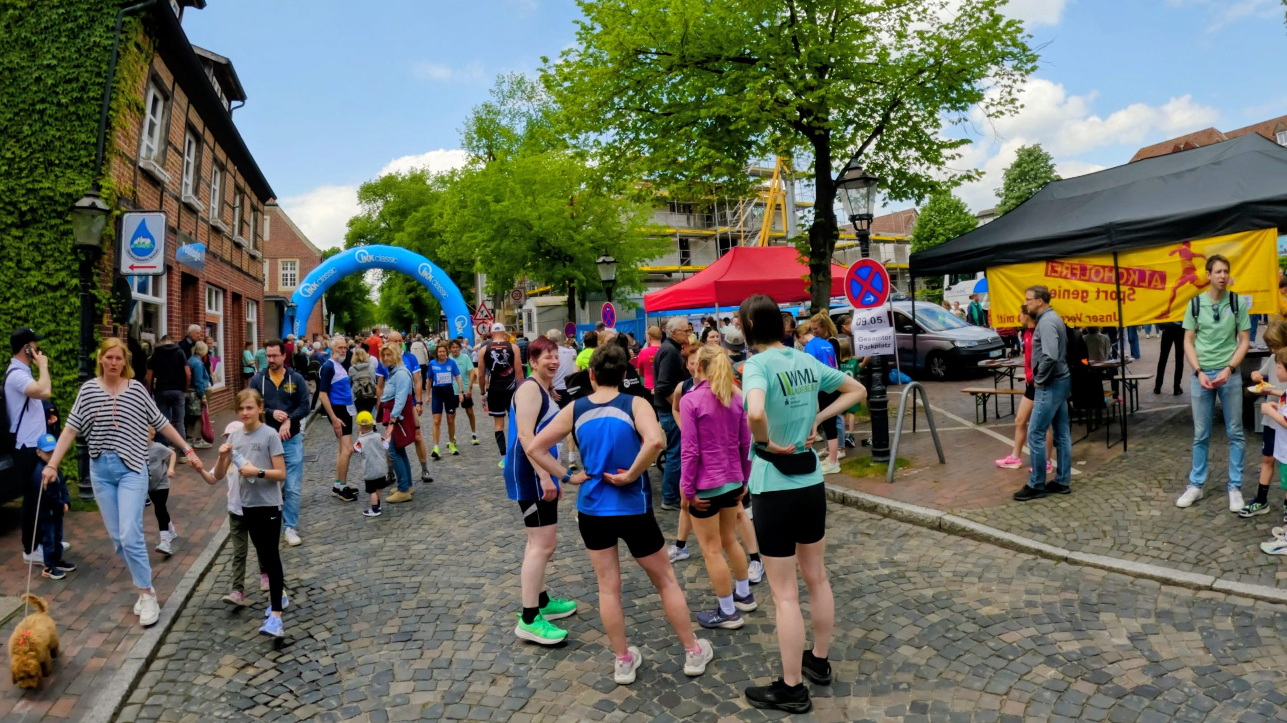 Crowd of runners and spectators on a cobblestone street during a community race, blue inflatable arch in background.