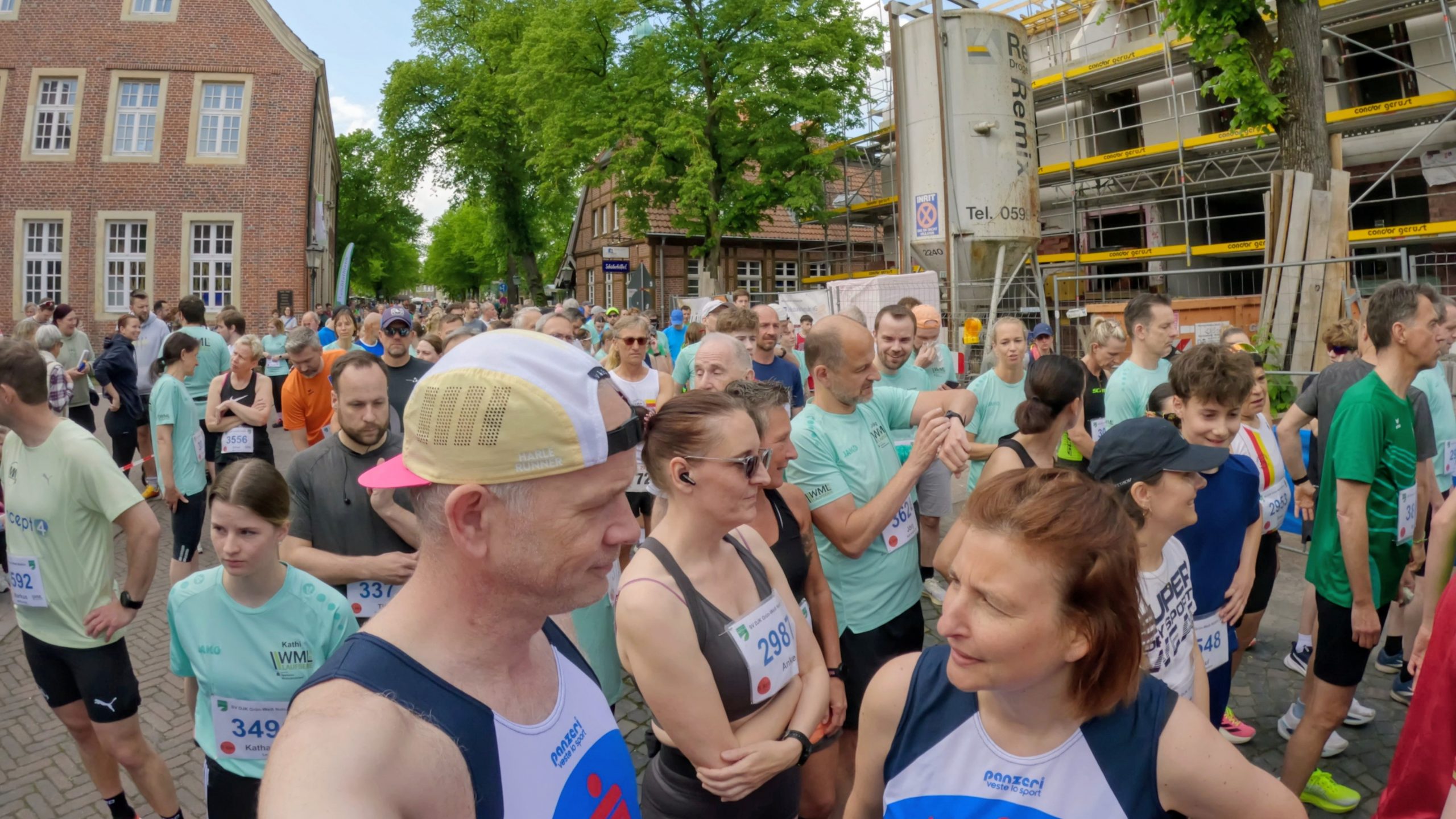 Large crowd of runners at a city street race start, with brick buildings to the left and a construction site on the right under green trees.