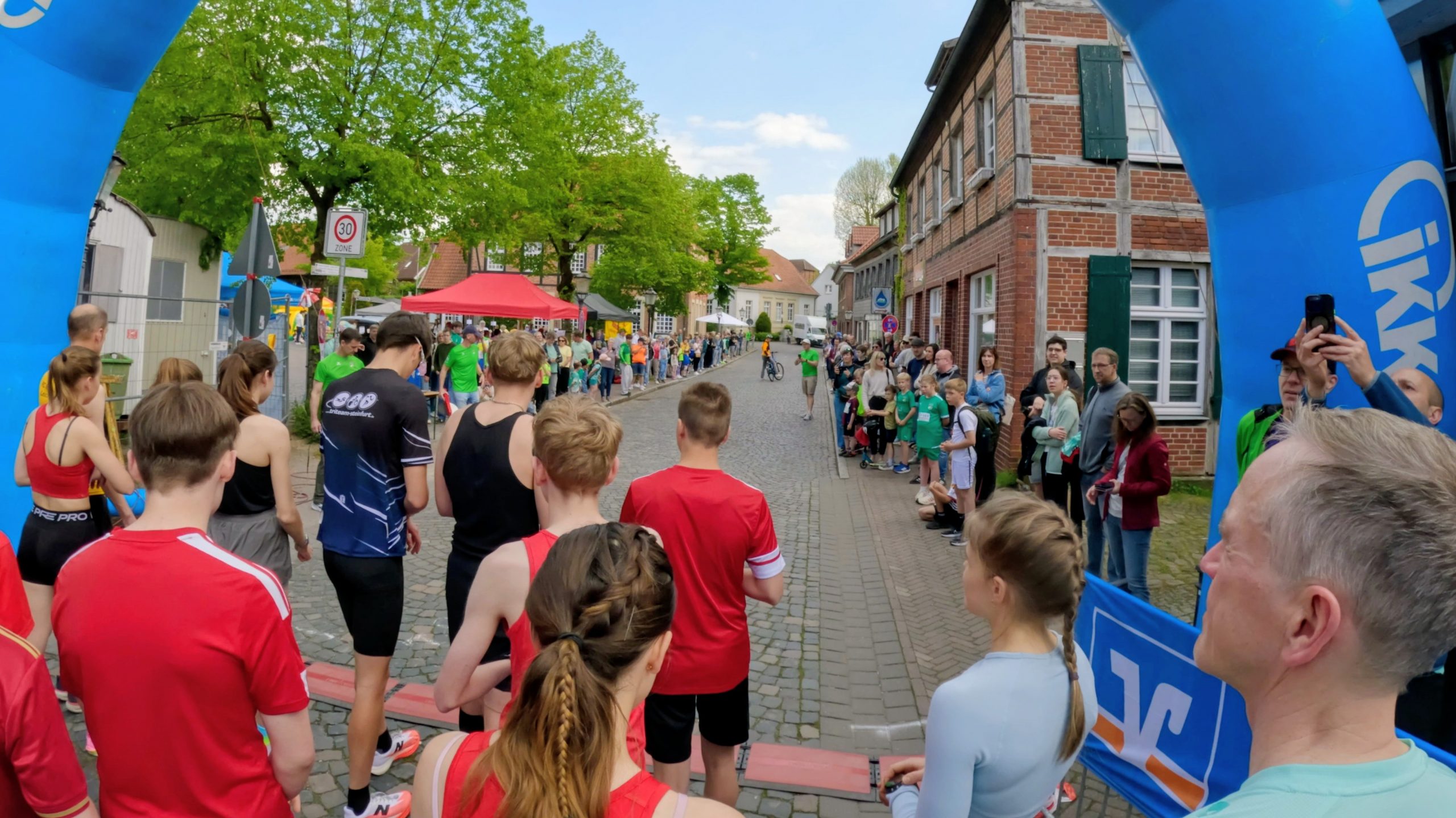 Crowd gathers along a cobblestone street as runners in red line up at the start under a large blue inflatable arch.