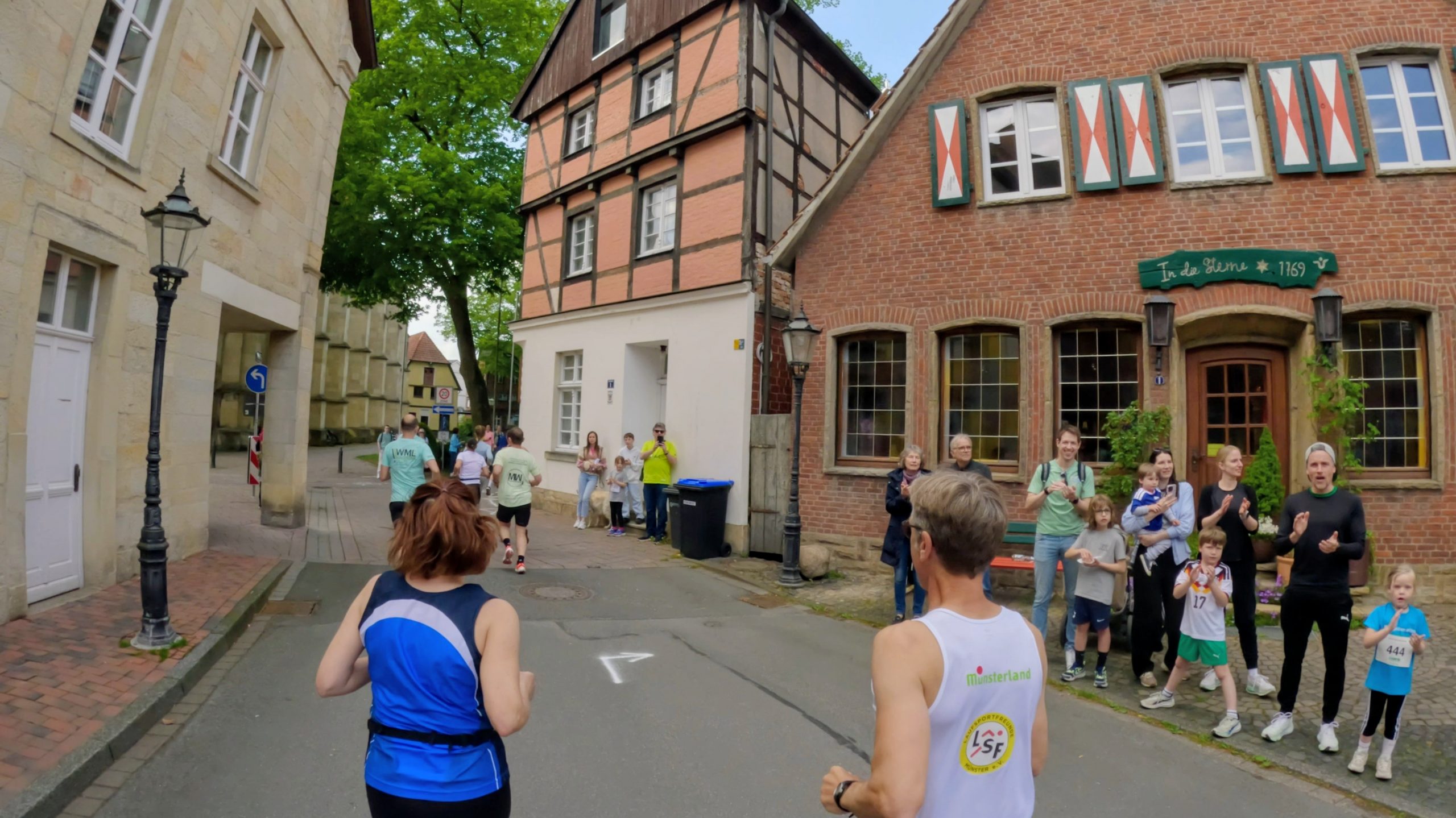 Runners participate in a street race past historic brick and timber-framed buildings with spectators cheering on the sidewalk.