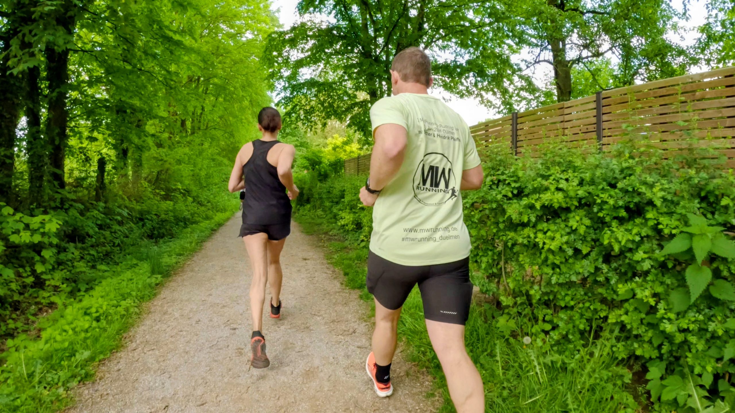 Two runners seen from behind on a tree-lined gravel path in a green park setting.