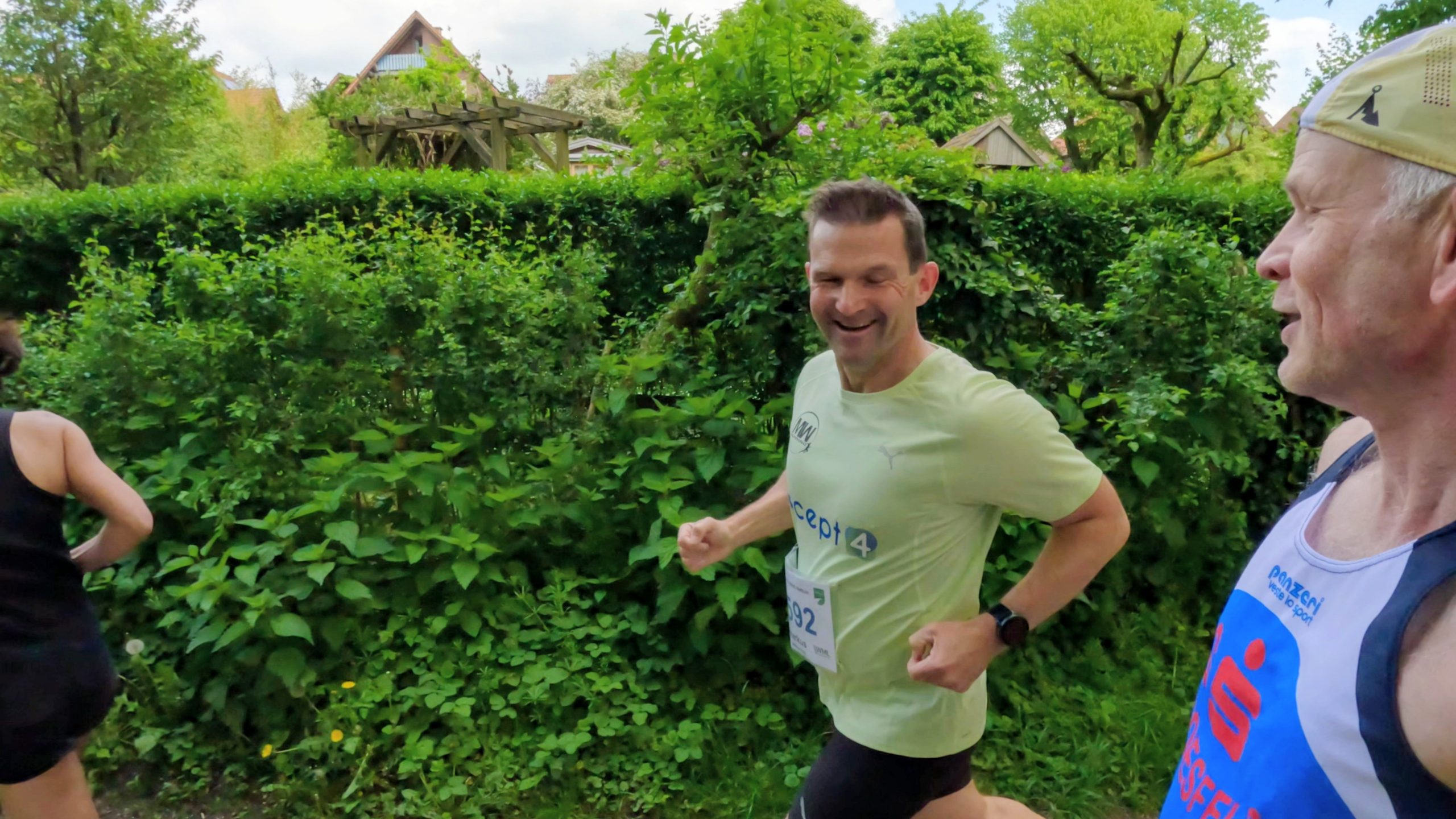 Three runners in a leafy park during a race, smiling and in motion along a path, with green hedges and trees surrounding them