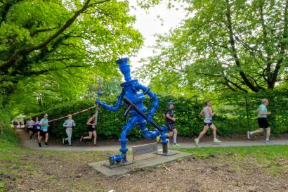 Blue industrial water pump sculpture in a park with runners in a race passing by behind it.