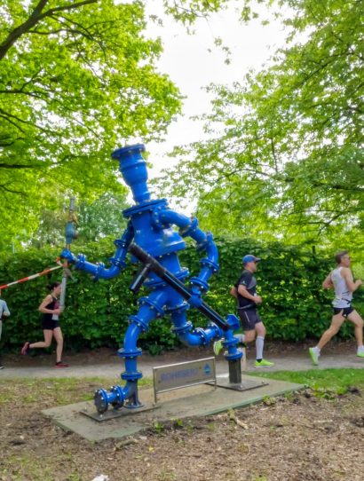 Blue industrial water pump sculpture in a park with runners in a race passing by behind it.