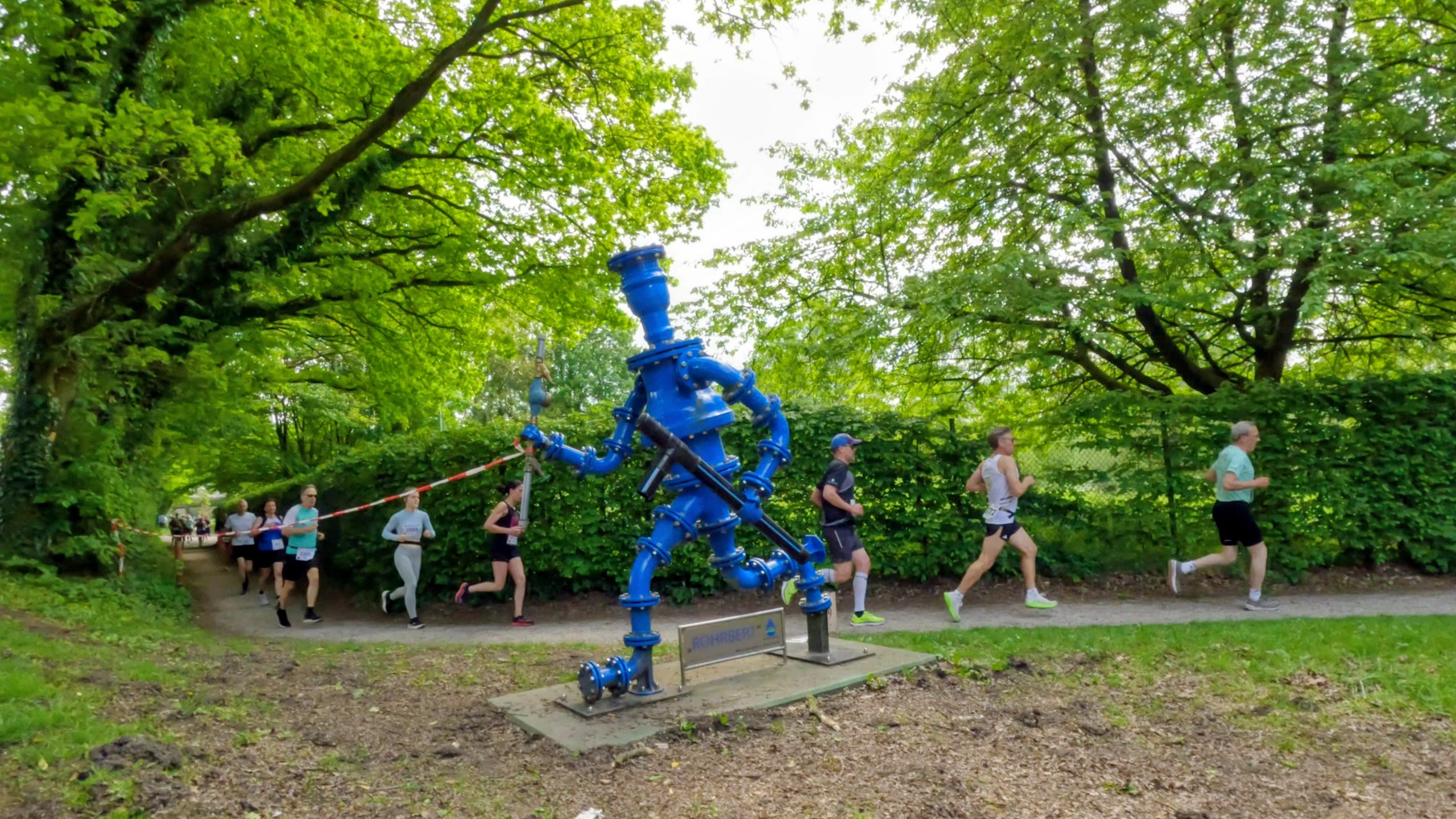 Blue industrial water pump sculpture in a park with runners in a race passing by behind it.