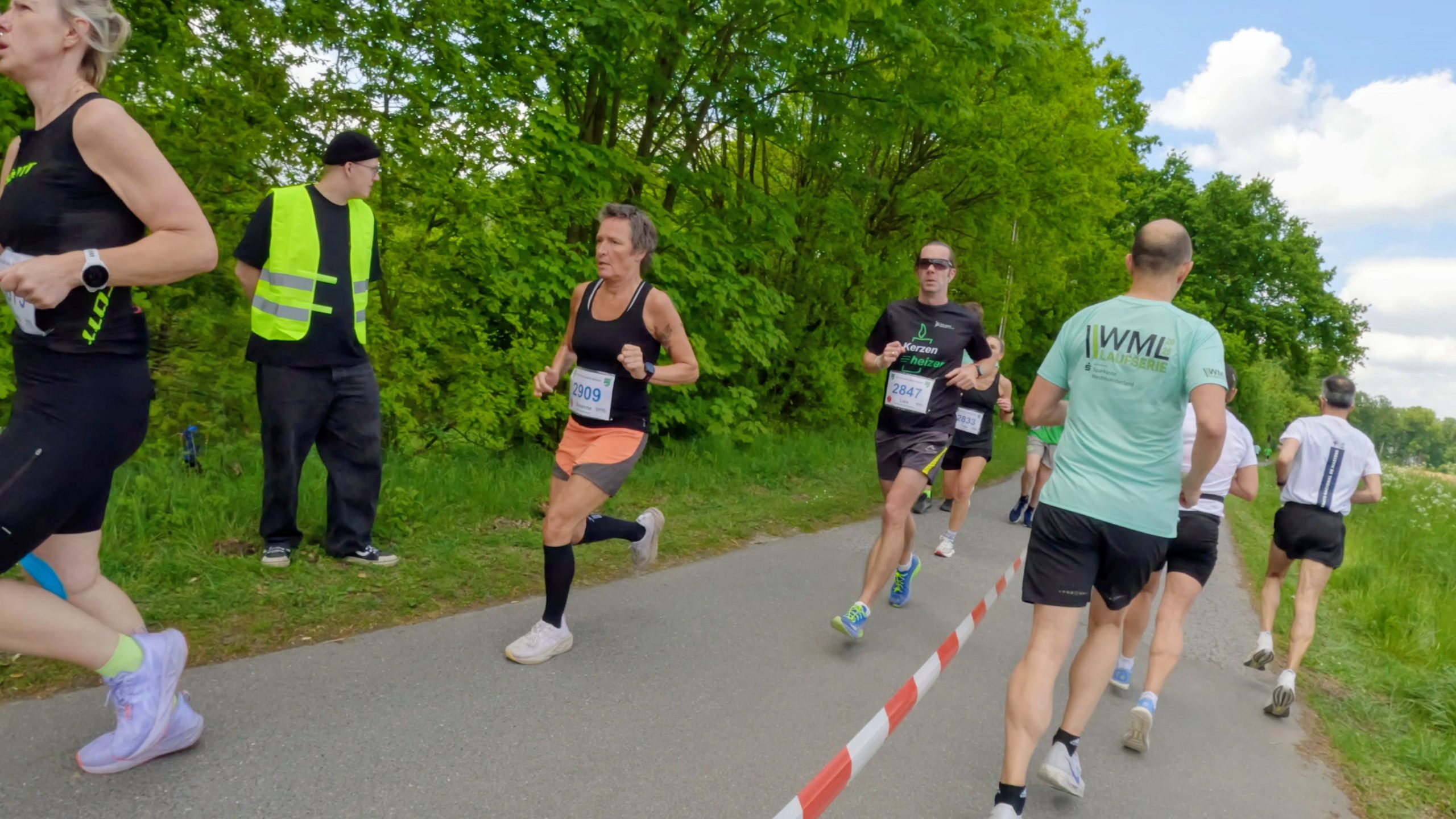 Runners in a road race pass by on a paved path lined with trees; spectators stand on grass beside the course, wearing numbered bibs and athletic gear.