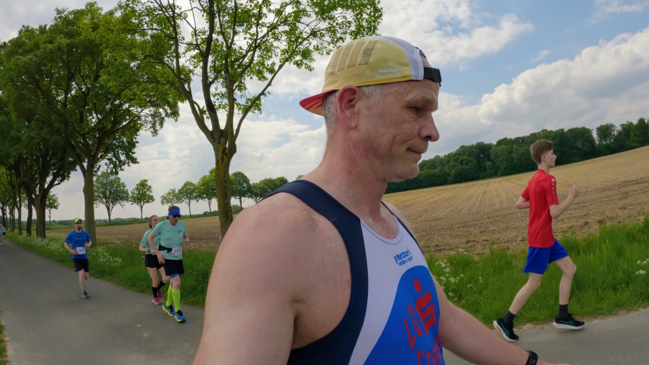 Runners participate in an outdoor race along a rural road, with a man in the foreground wearing a cap and race bibs visible behind him.