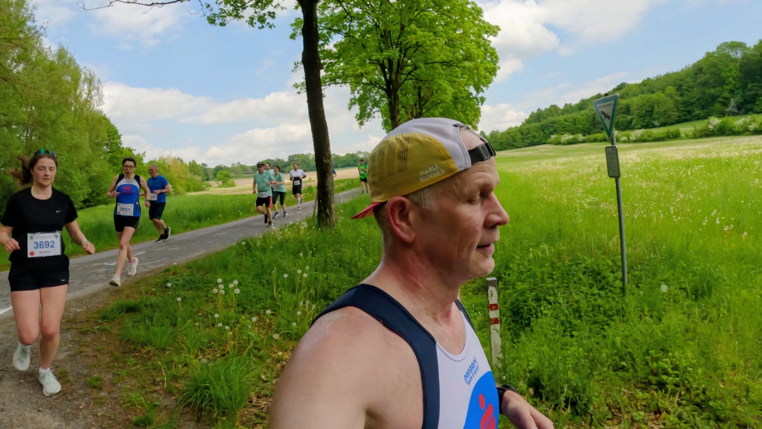 Runners in a countryside race along a narrow path, the foreground man wearing a beige cap and sleeveless top, with green fields and trees on a sunny day.