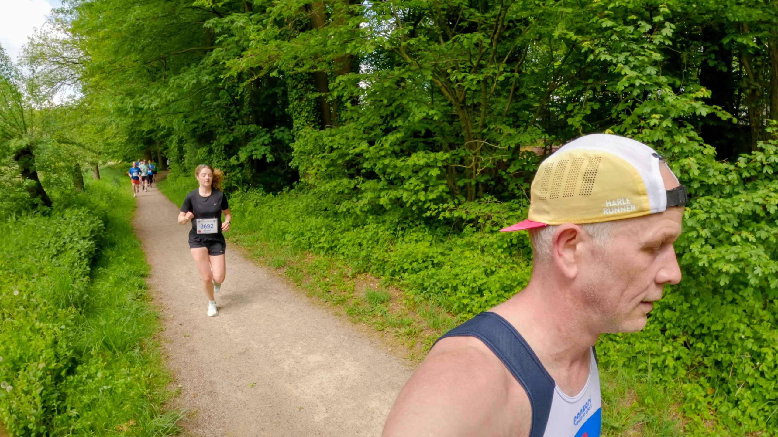 Runners on a dirt path through a green forest, foreground man wearing a cap and sleeveless shirt attack the trail.