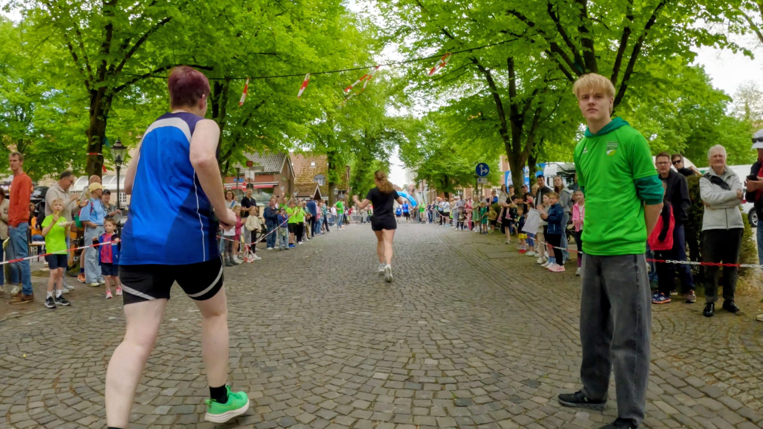 Runners compete in a tree-lined cobblestone street with cheering spectators on both sides during a race. In the foreground, a runner in blue passes by while a man in a green shirt stands to the right.