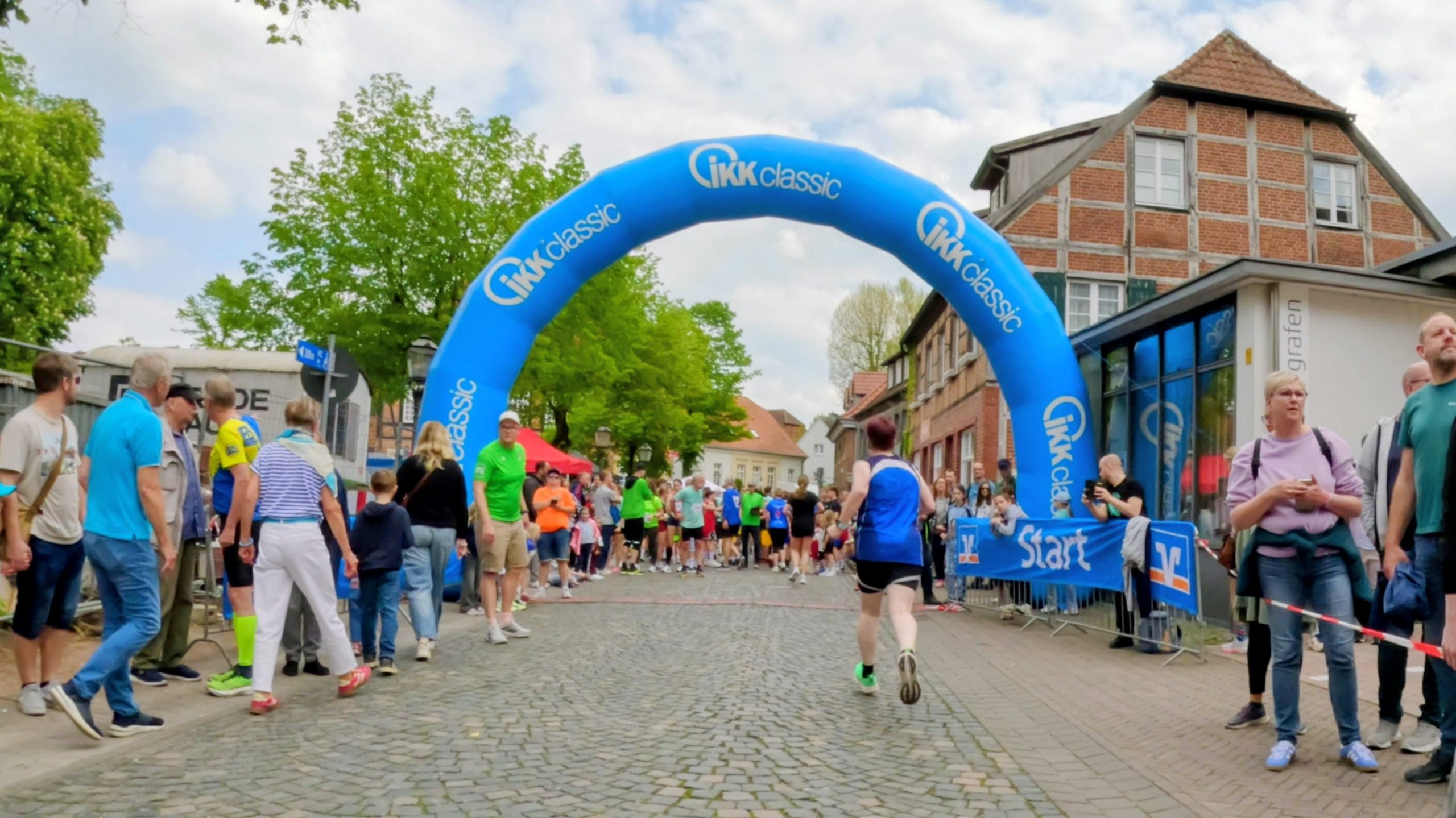 Crowd gathers at a race start under a blue inflatable arch reading IKK classic on a cobblestone street.
