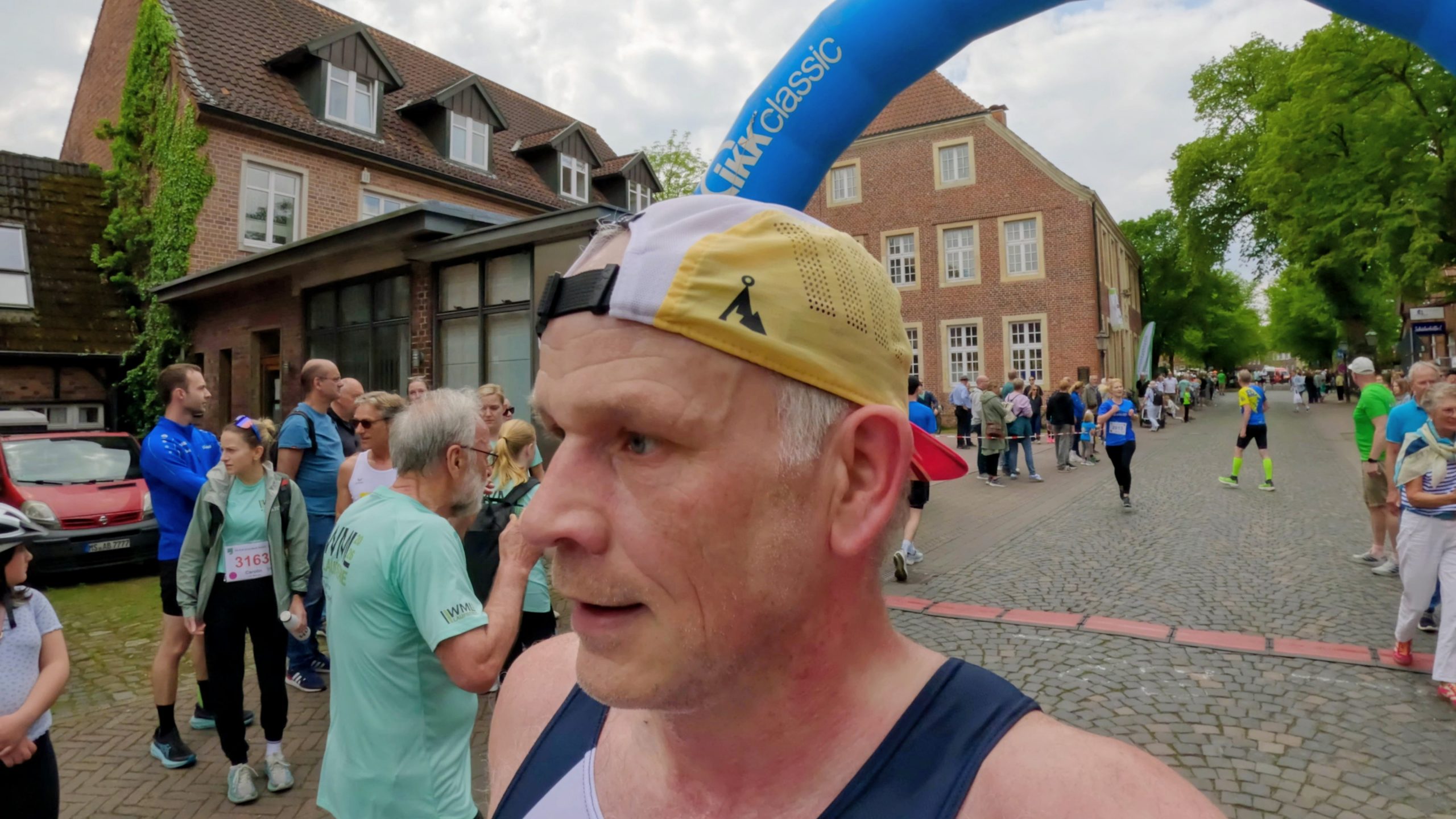 Close-up of a runner wearing a beige headband and navy singlet at a city street race, with spectators and brick buildings in the background.