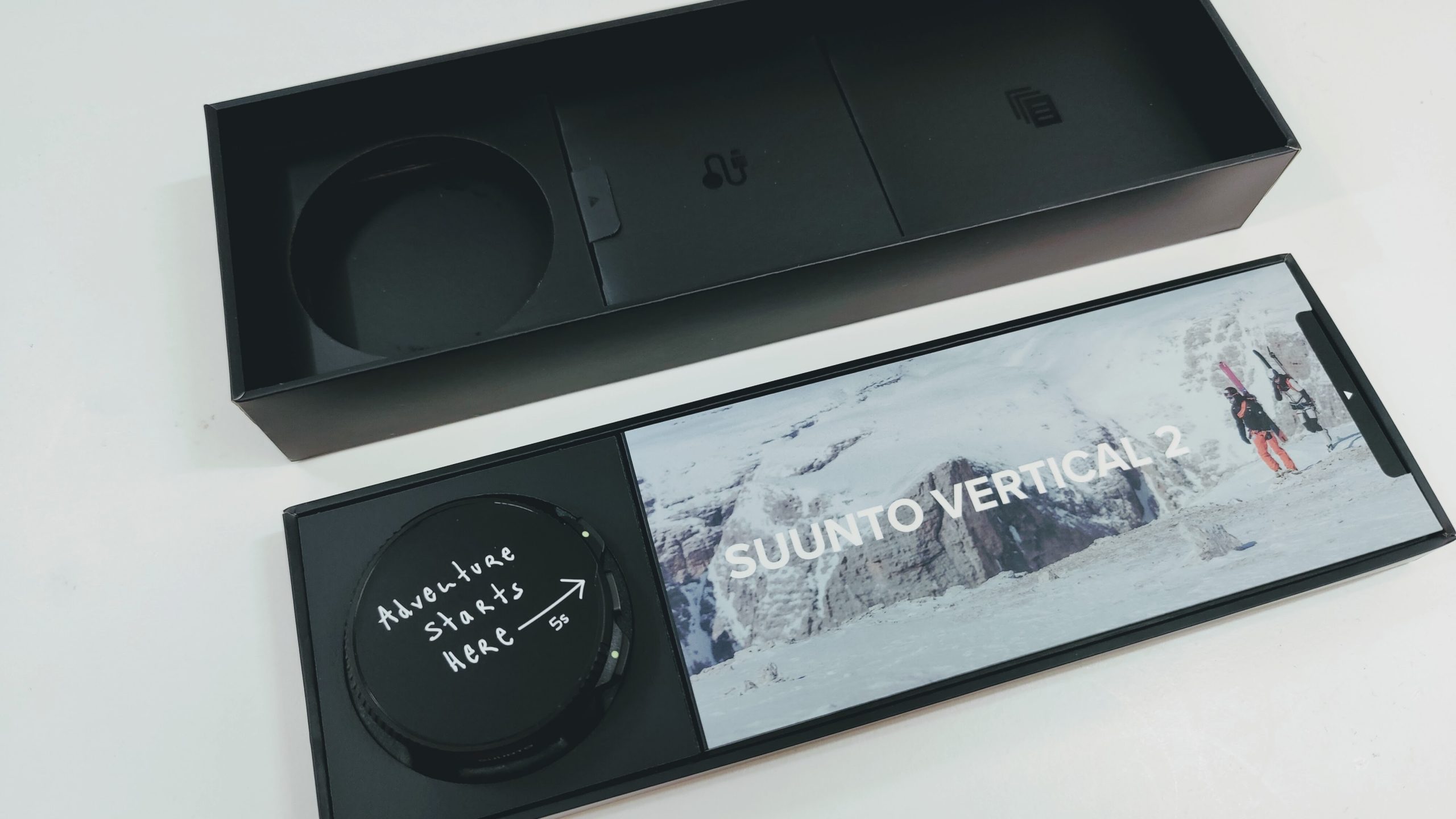 Open black packaging for a Suunto Vertical 2, featuring a circular left compartment and a right insert with a snowy mountain photo and the text 'SUUNTO VERTICAL 2'. The left lower section shows a round dial with the message 'Adventure starts here'.