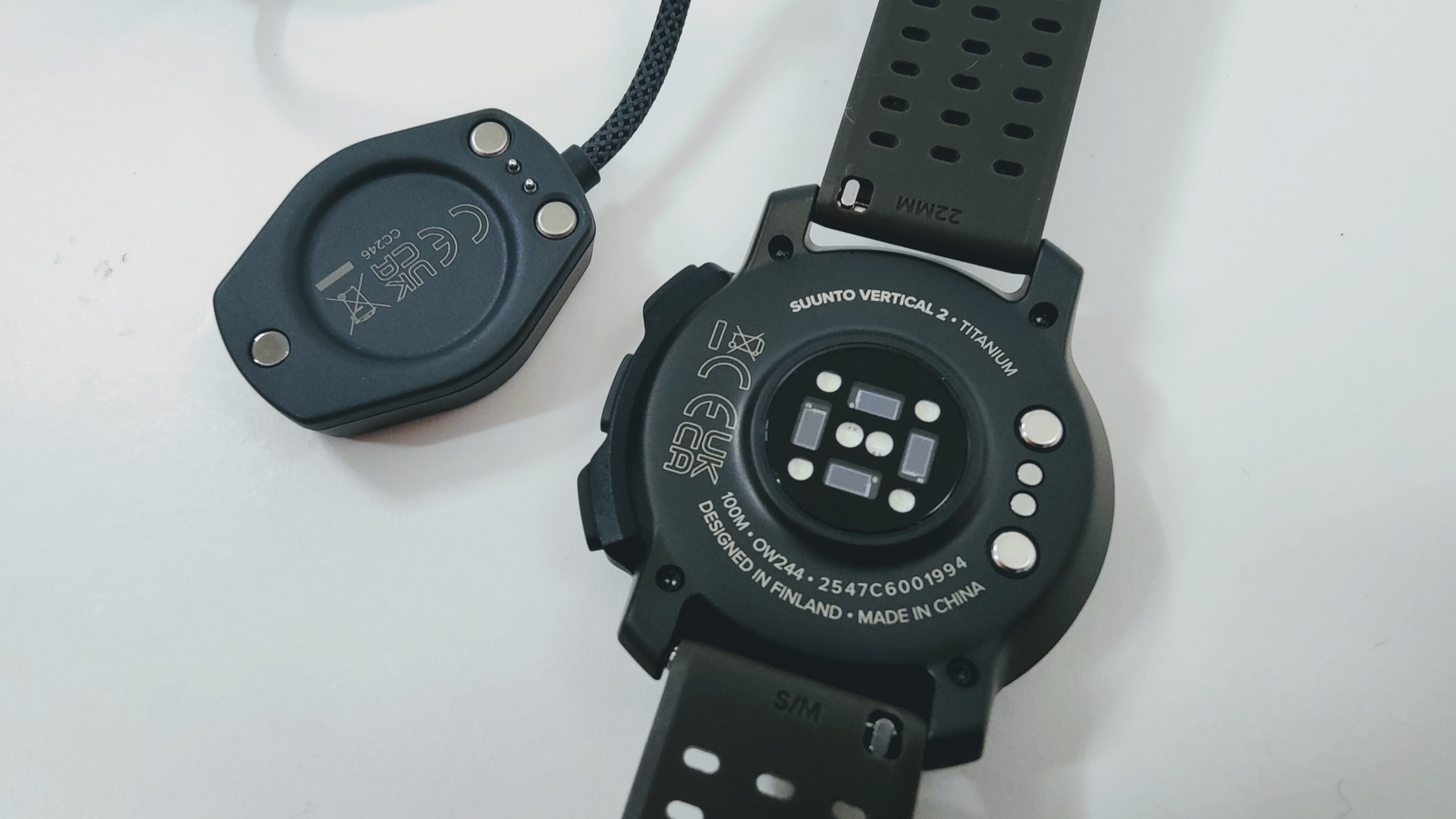 Close-up of a black Suunto Vertical 2 Titanium smartwatch back with sensor array and a nearby charging cradle.