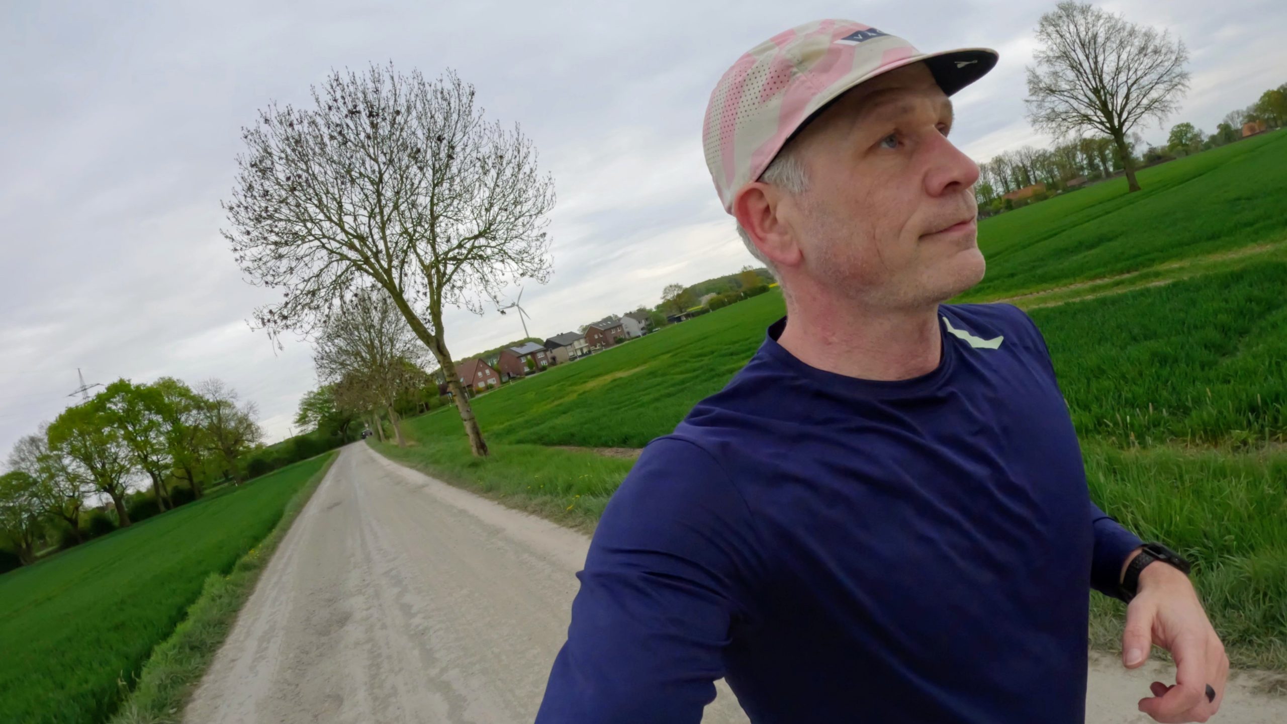 Man in a navy shirt and pink cap jogging along a rural road with green fields and trees in the background.