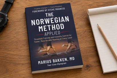 Navy book titled The Norwegian Method Applied on a wooden desk beside a notebook and a black sports watch left of the book