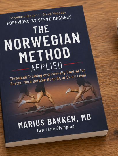 Navy book titled The Norwegian Method Applied on a wooden desk beside a notebook and a black sports watch left of the book