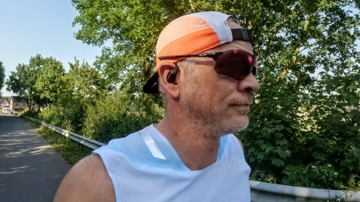 Man jogging on a sunny, tree-lined road wearing a white sleeveless shirt, orange/white cap worn backwards, sunglasses, and wireless earbuds.