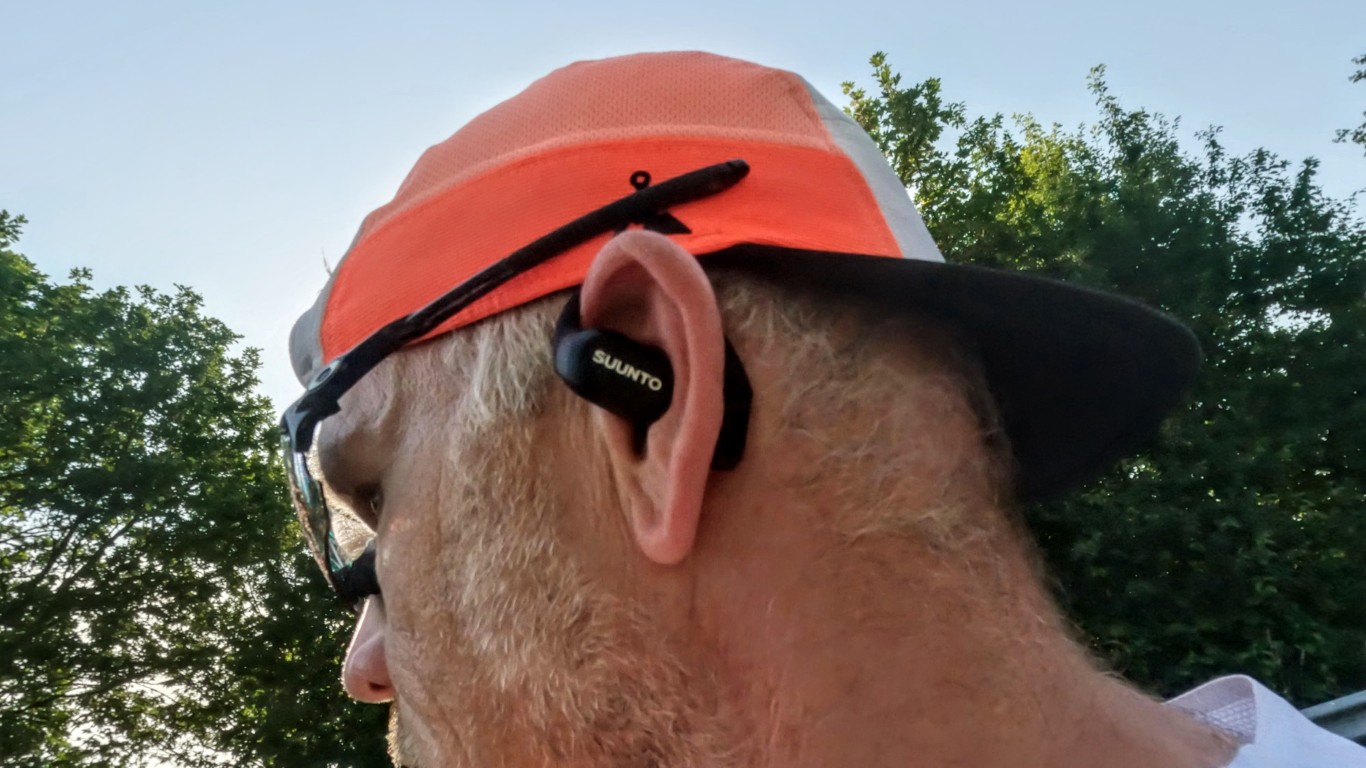 Close-up of a person wearing an orange cap and black Suunto earbud outdoors with trees in the background.