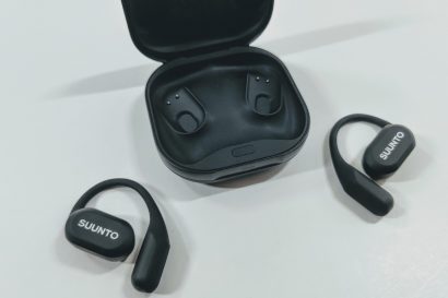 Wireless black earbuds with charging case open; Suunto logo on earbuds, one earbud outside case and one nearby on a light surface.