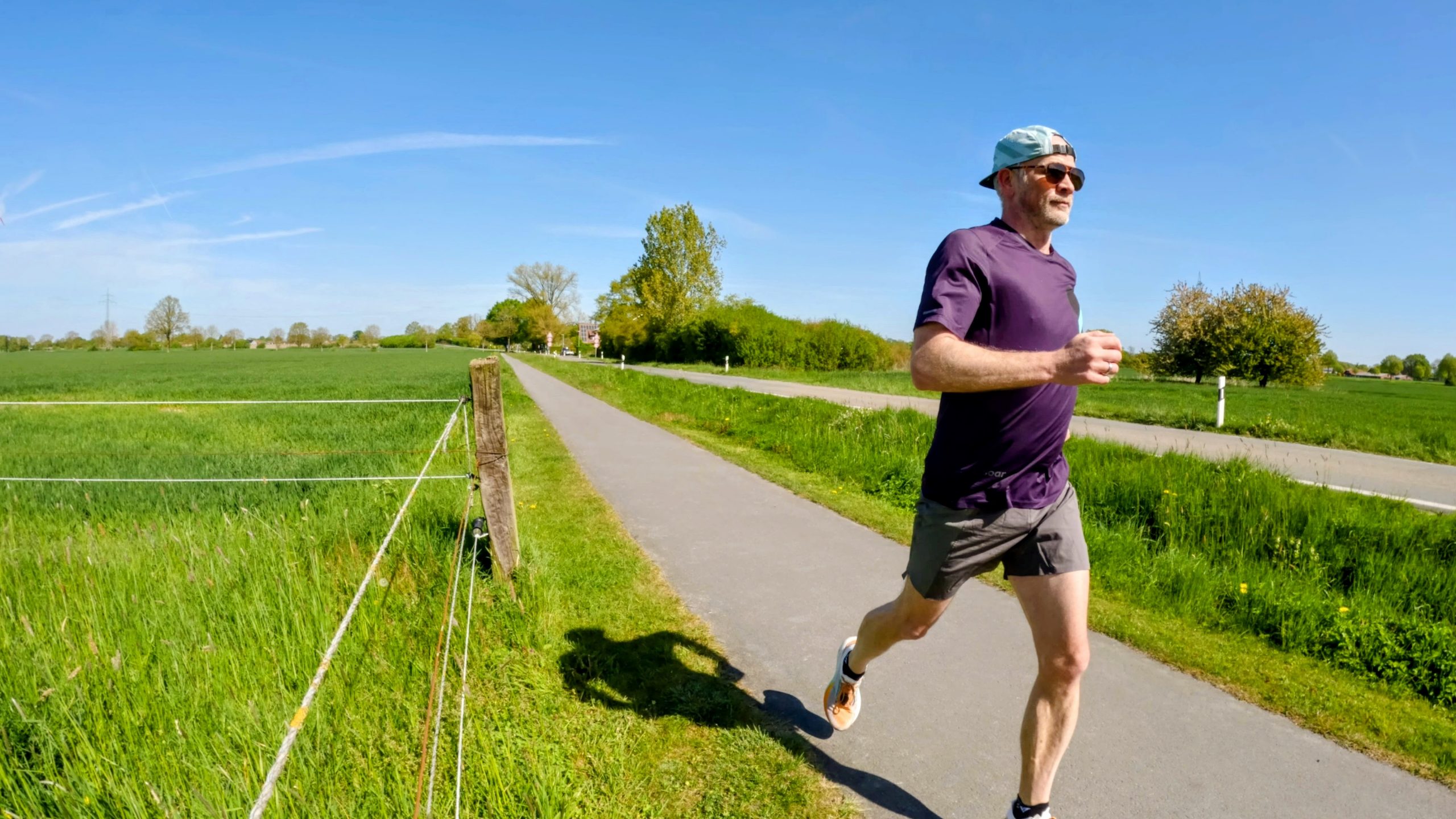 Man jogging on a rural paved path, wearing a purple shirt, gray shorts, sunglasses, and a backward cap, with green fields and blue sky.