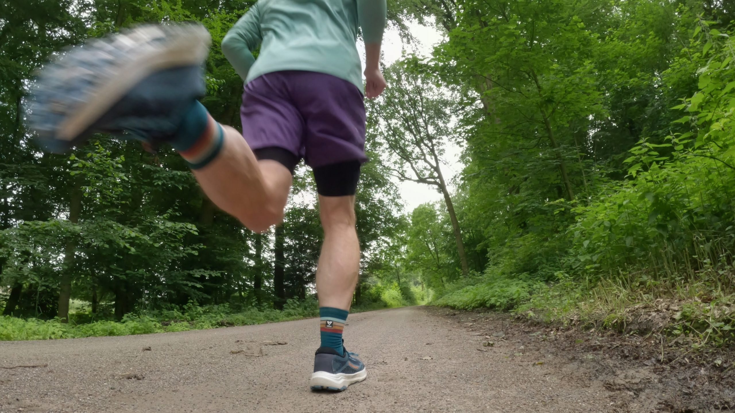 Low-angle shot of a person running on a forest path, wearing teal top and purple shorts with trees in the background