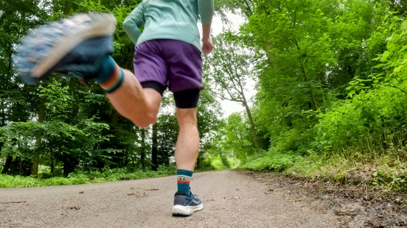 Runner seen from behind mid-stride on a forest trail, wearing teal jacket, purple shorts, and teal shoes amidst green trees.