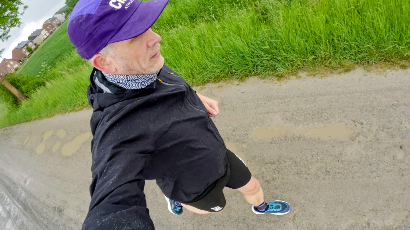 Man wearing a purple cap and black jacket running along a muddy dirt path with tall green grass and houses in the background, taken from an over-the-shoulder selfie angle.