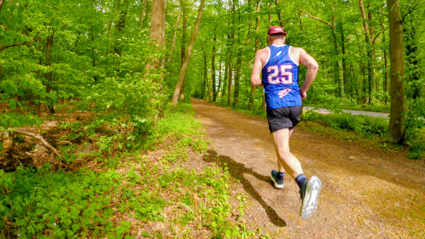 Runner wearing a blue jersey with the number 25 jogging along a dirt forest path, surrounded by green trees (back view).