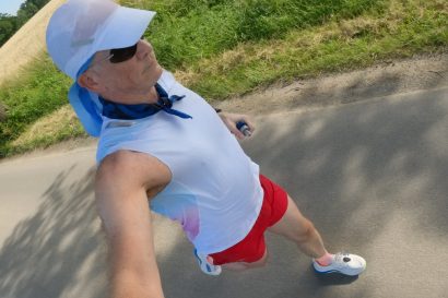 Man wearing a white cap with a blue scarf and sunglasses running on a paved path, grass and trees to the side.