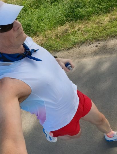 Man wearing a white cap with a blue scarf and sunglasses running on a paved path, grass and trees to the side.