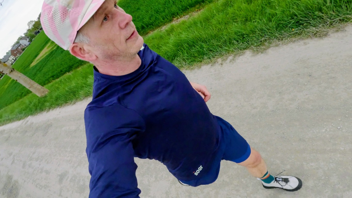 Man jogging on a gravel path in a park, wearing a navy shirt, blue shorts, white-pink cap, and running shoes.