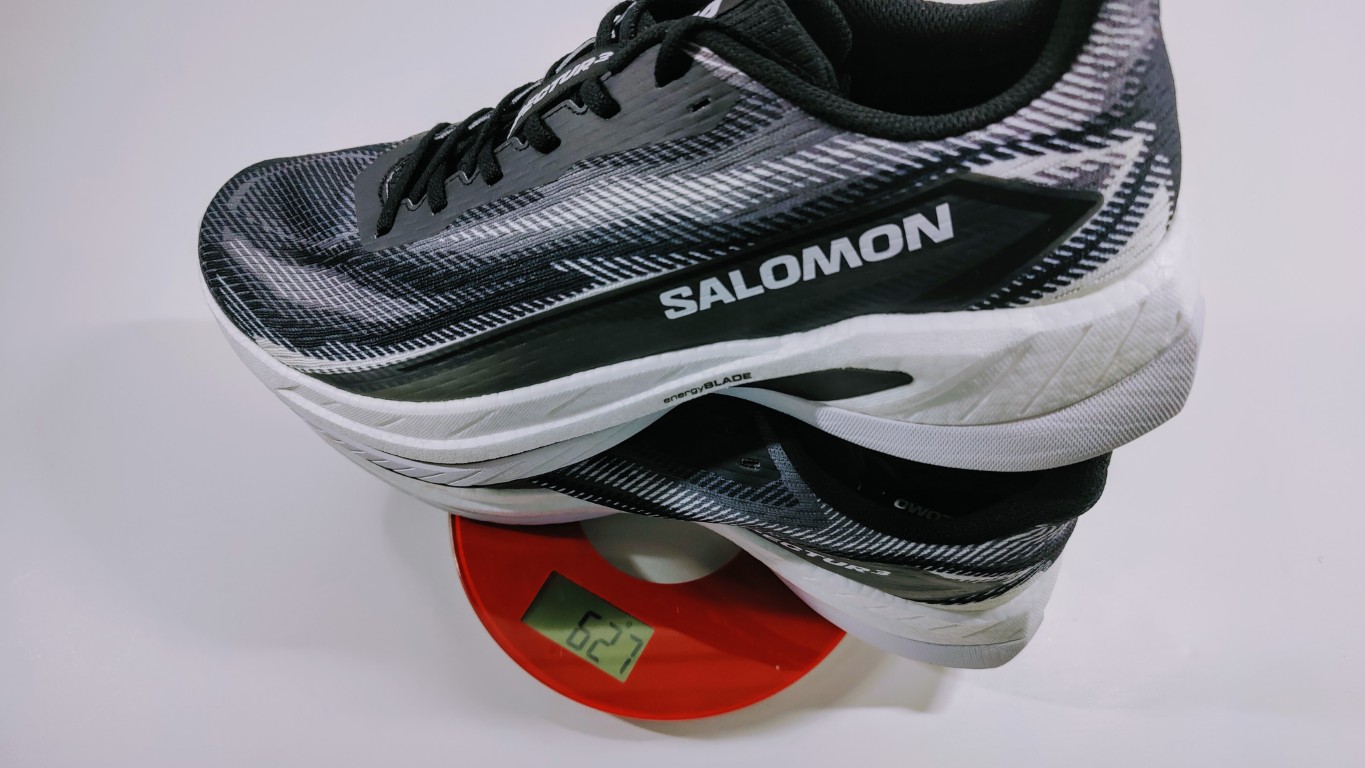 Pair of Salomon running shoes stacked over a red digital scale displaying about 627 g.