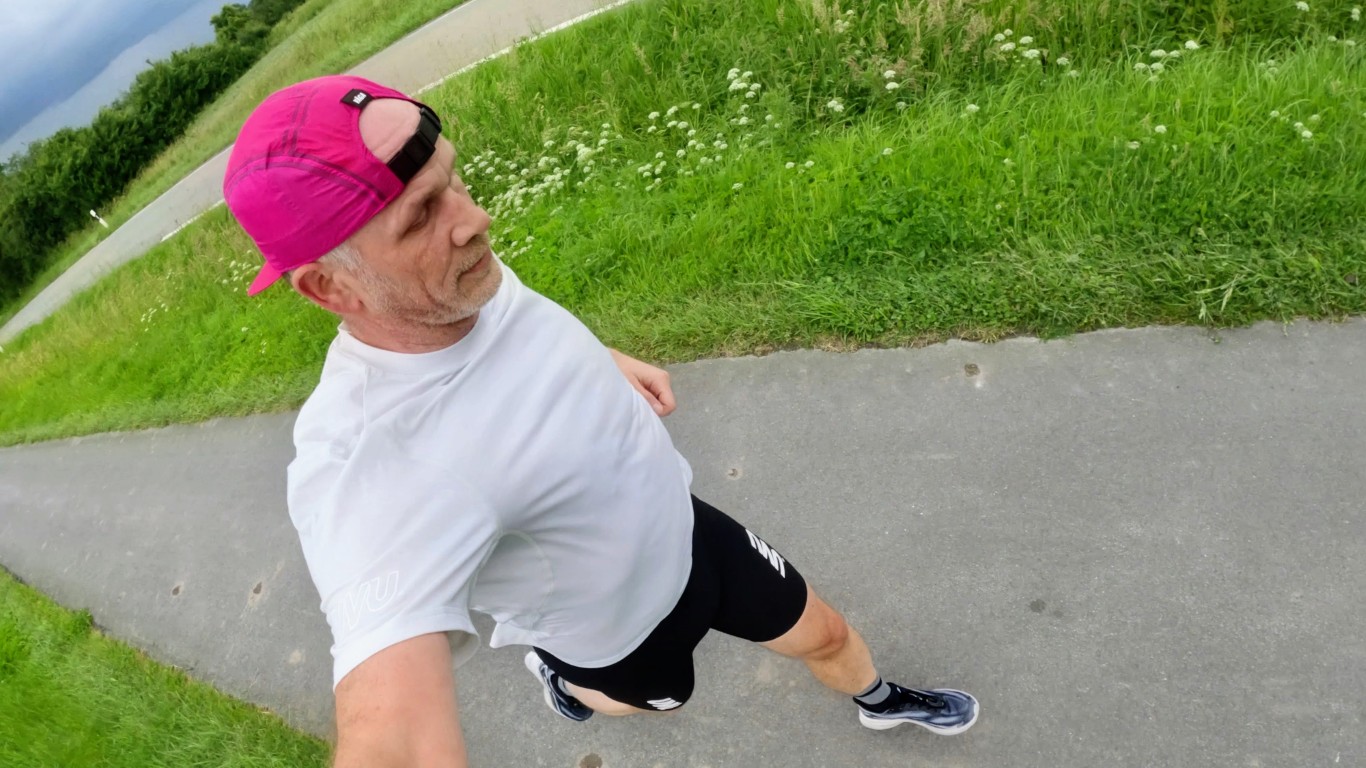 Man in a white T-shirt and black shorts runs along a paved path in a park, wearing a pink cap backward.