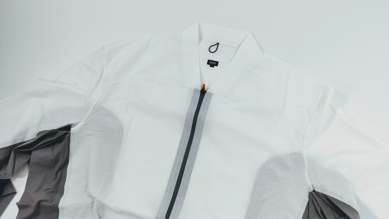 White athletic jacket laid flat with a vertical black zipper running down the center and gray sleeves. The brand tag is visible near the collar.