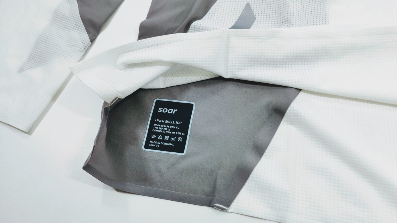 Close-up of a white textured fabric with a grey panel showing a black label that reads 'soar' Linen Shell Top on the garment surface.