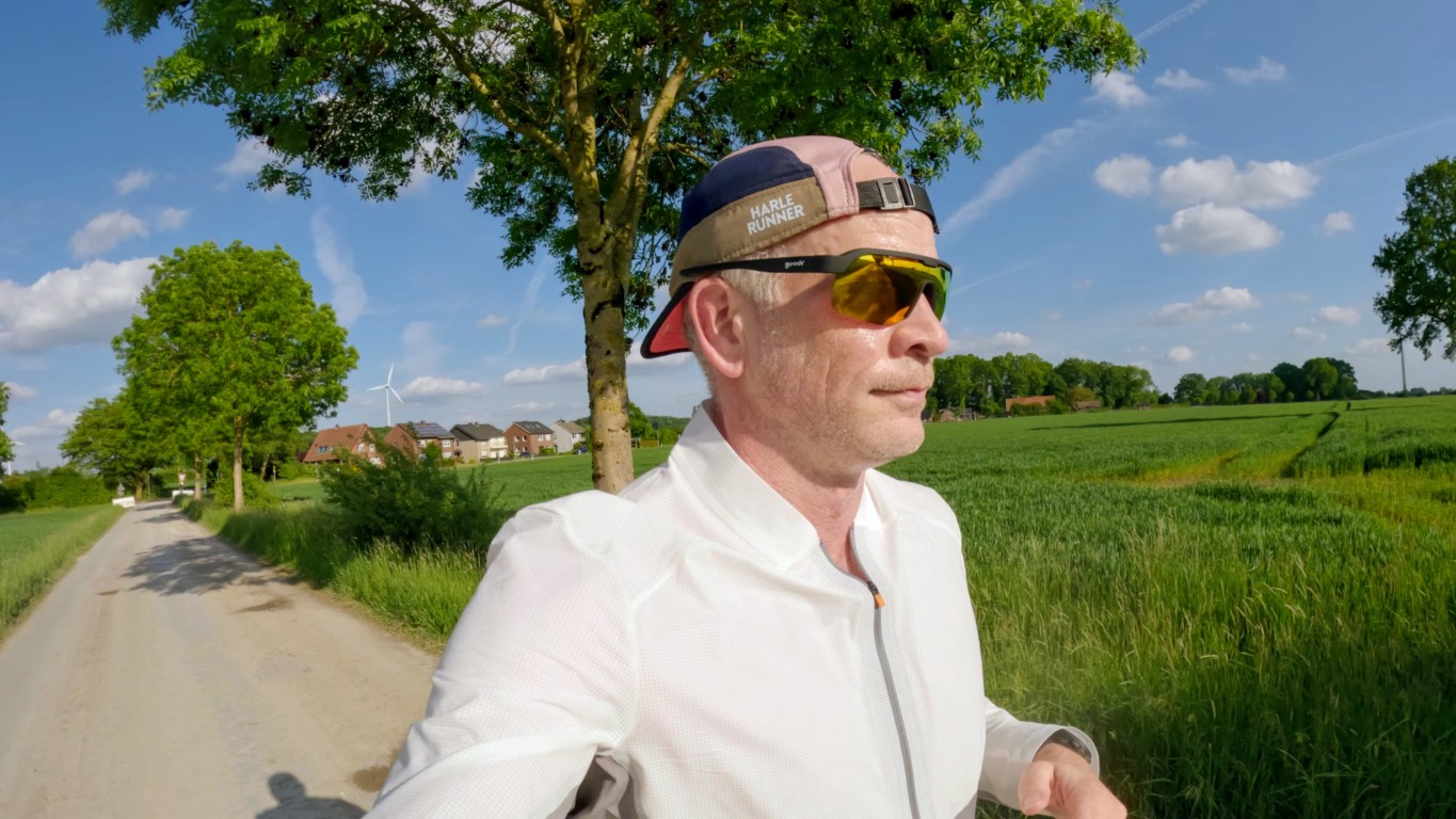 Runner in a white jacket and orange-tinted sunglasses runs along a sunny rural dirt road with green fields and a wind turbine in the distance.
