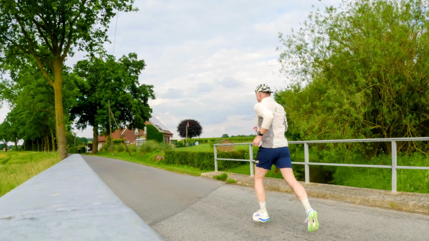 Runner wearing a light jacket and blue shorts jogging along a rural road beside a metal railing, with trees and a house in the distance.