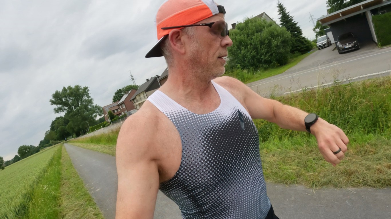 Man jogging along a paved path beside grassy fields, wearing an orange visor cap, sunglasses, and a patterned sleeveless running shirt.